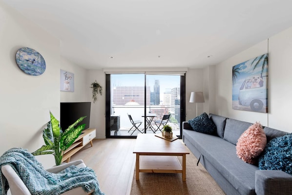 A modern living room with a sofa, coffee table, and a TV on a stand. There is a balcony with a view of the city.