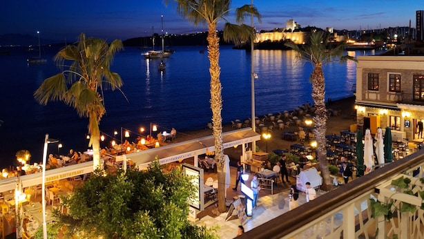 A seaside terrace with outdoor seating, palm trees, and a view of the sea and a castle in the background. - SAND GARDEN (Bodrum)
