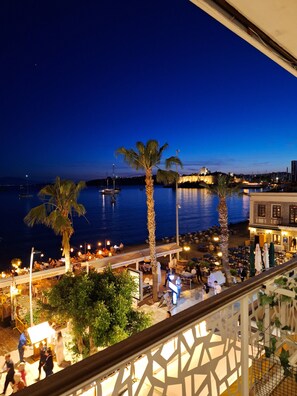 A seaside terrace with outdoor seating, palm trees, and a view of the sea and a castle in the background. - SAND GARDEN (Bodrum)
