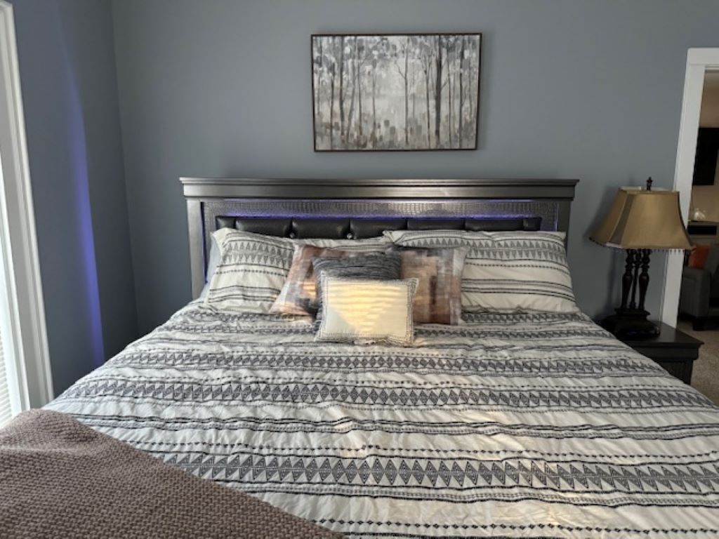 Une chambre à coucher avec une couvre-lit à motifs, une tête de lit dotée d’un éclairage intégré, une œuvre d’art encadrée au mur et une table de chevet avec une lampe.