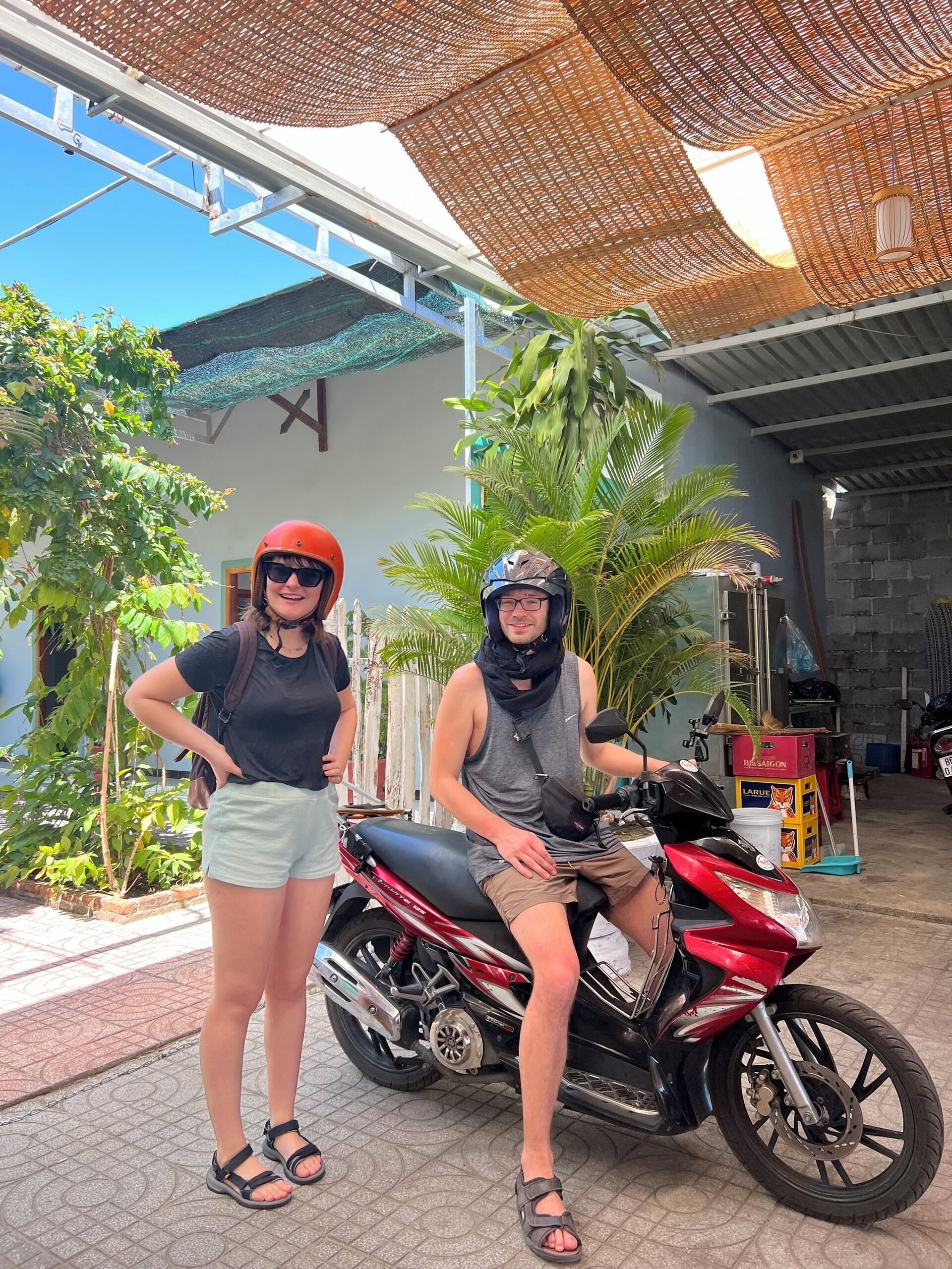 Two people standing next to a motorcycle in a covered outdoor area with plants and a building in the background.
