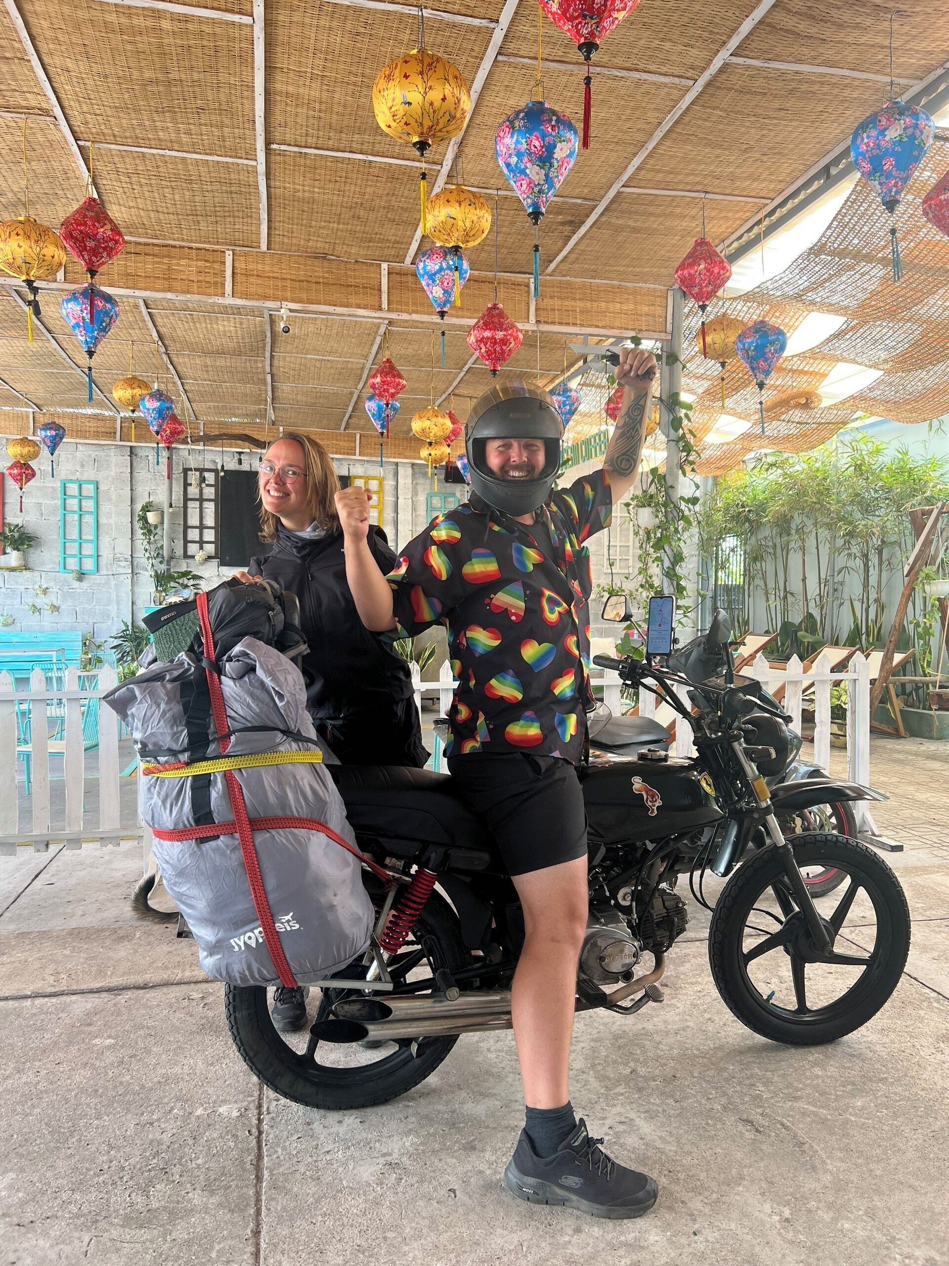 Una motocicletta con un passeggero, entrambi con caschi, parcheggiata sotto un tetto di paglia decorato con lanterne colorate.