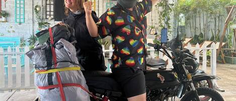 Una motocicletta con un passeggero, entrambi con caschi, parcheggiata sotto un tetto di paglia decorato con lanterne colorate.