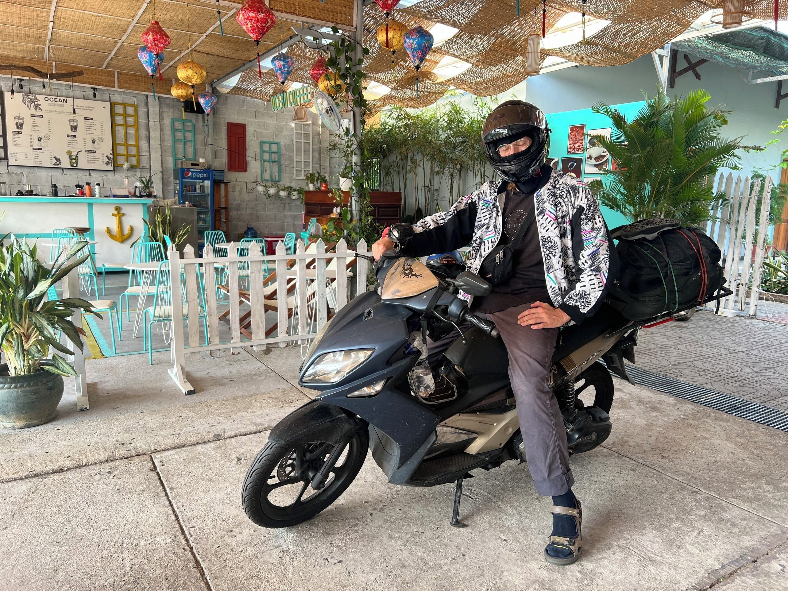 Una persona sentada en una motocicleta, con casco, una chaqueta estampada y una mochila.