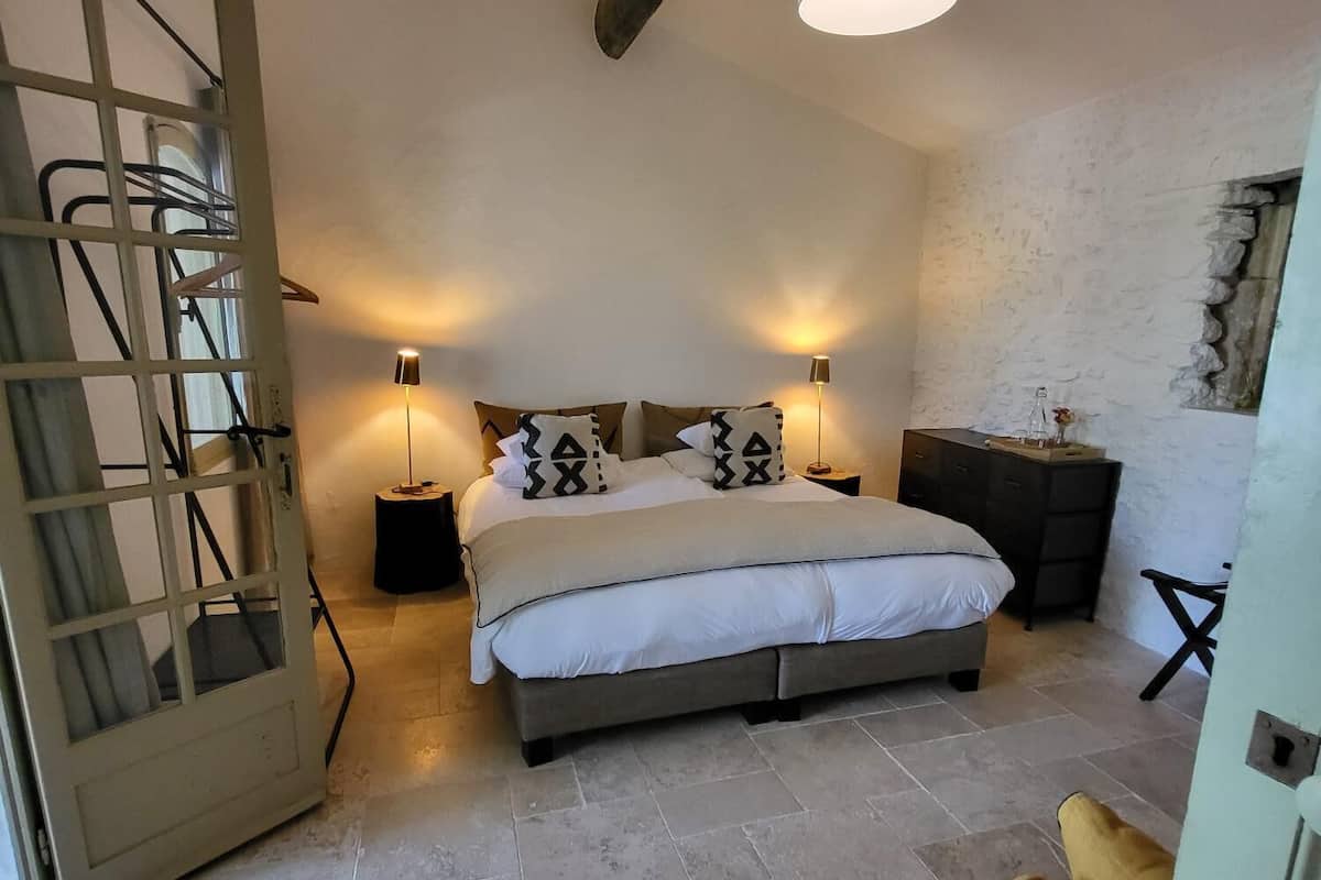 A bedroom with a bed, two bedside tables, a hanging light fixture, a wooden ladder, and a stone wall.