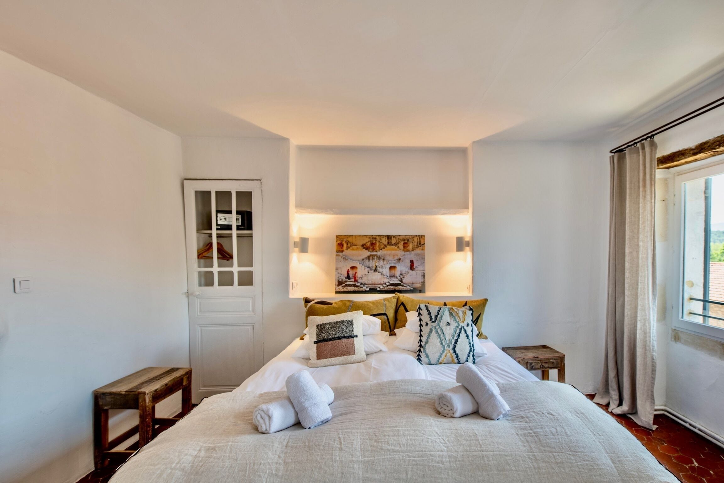 A neatly made bed with towels, a wooden bench, a door with glass panels, and a painting above the bed.