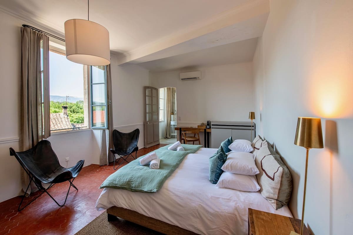 A bedroom with a large bed, a desk, a chair, and a view of greenery outside.