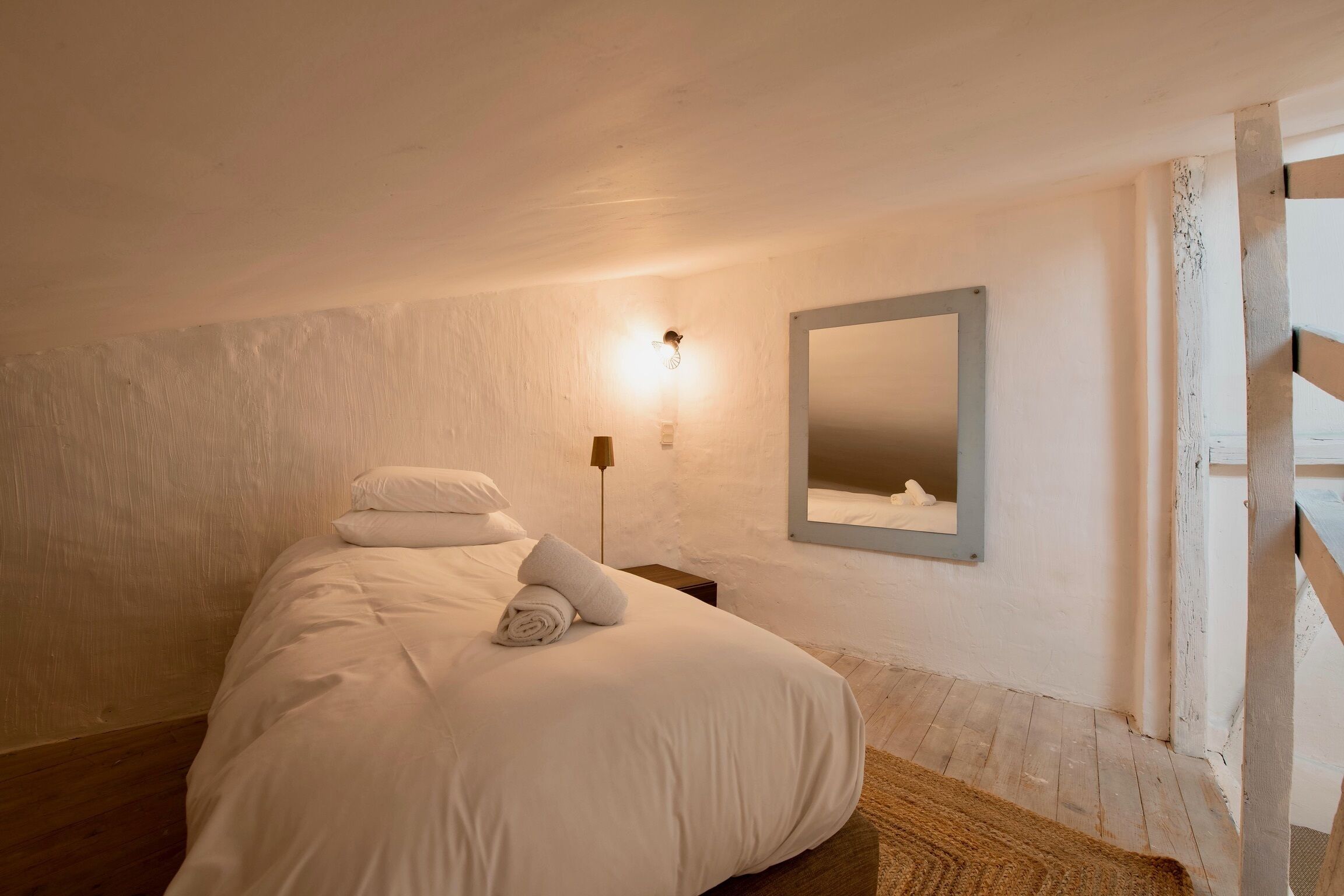 Un dormitorio con cama, mesita de noche, espejo en la pared y suelo de madera.