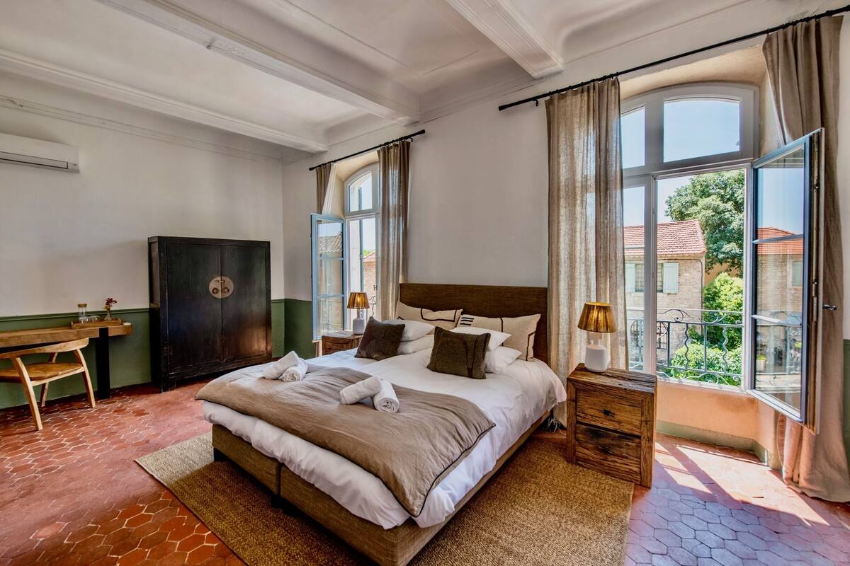 A bedroom with a large bed, a wooden desk, a wardrobe, and a view of the outside.