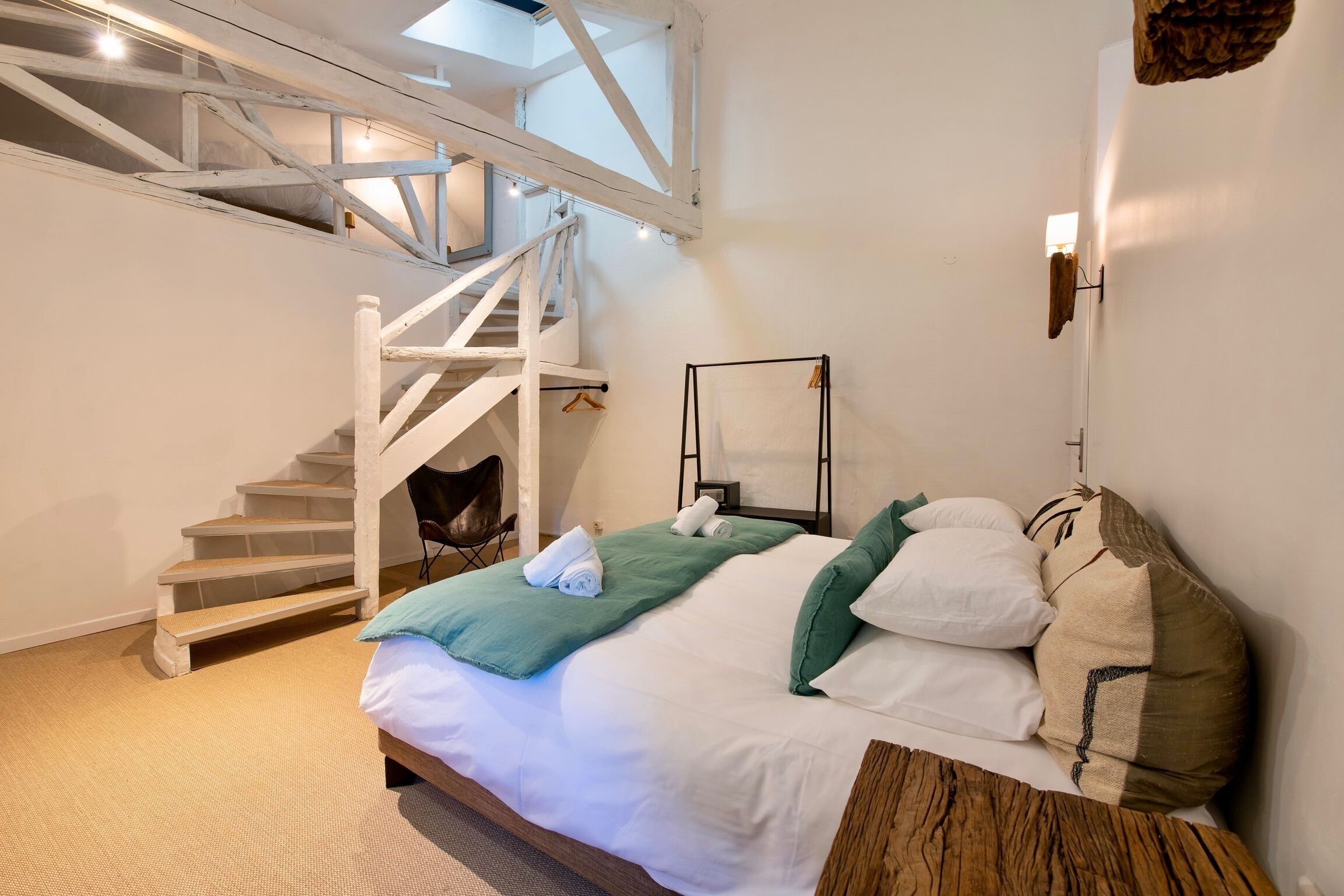 A bedroom with a bed, a staircase, a hanging light fixture, and a wall-mounted mirror.