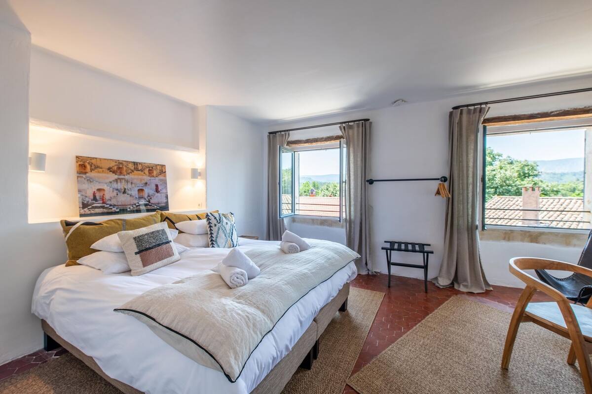 A neatly made bed with white linens and multiple pillows, a painting on the wall, a bench, and a view of greenery through the window.