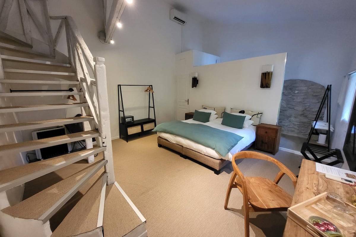 A bedroom with a bed, a wooden chair, a desk, a hanging chair, a staircase, and a stone wall.
