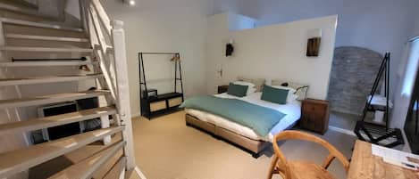 Une chambre à coucher avec un lit, une chaise en bois, un bureau, un fauteuil suspendu, un escalier et un mur en pierre.