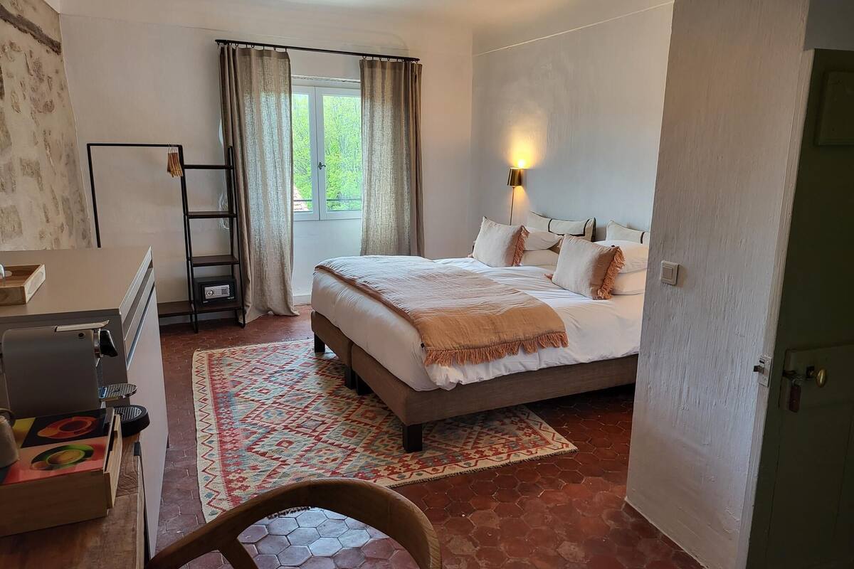 A bedroom with a large bed, a desk with a computer, a chair, a window with curtains, and a view of greenery outside.