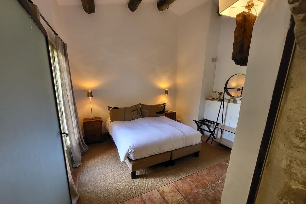 A bedroom with a bed, two bedside tables, a chair, a lamp, and a wall-mounted shelf.