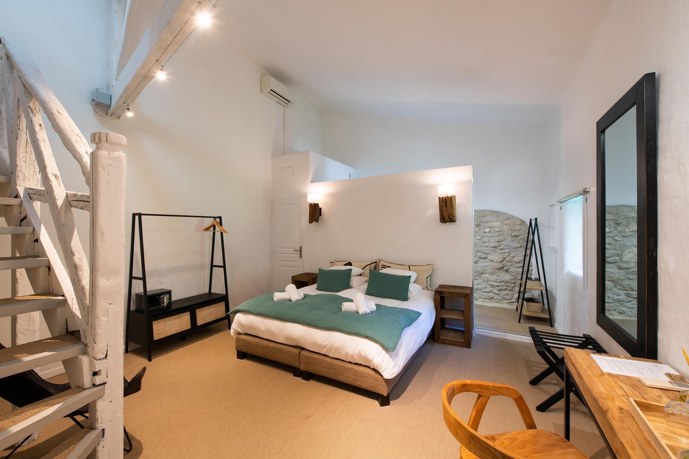 A bedroom with a bed, a desk, a chair, a ladder, a hanging light, and a stone wall.