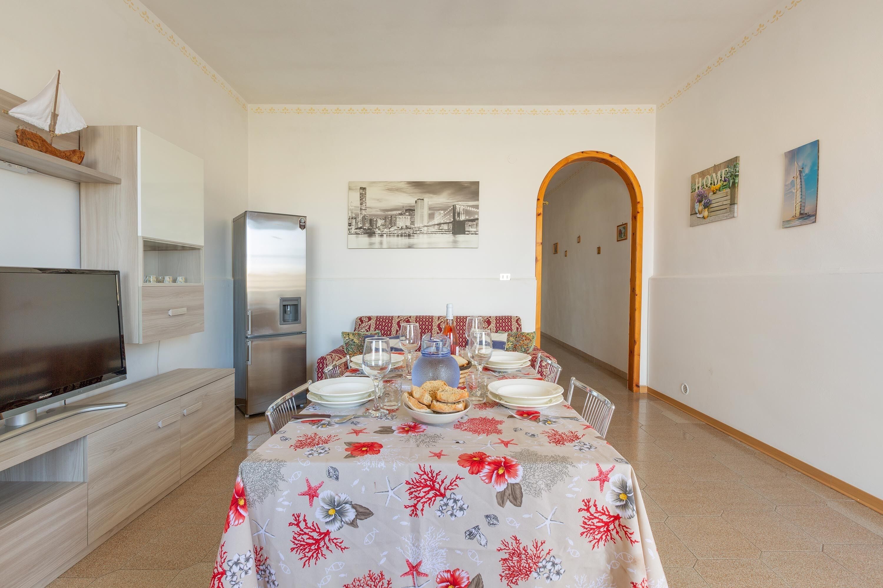 A dining area with a table set for four, a refrigerator, a television, and a decorative sailboat on a shelf.