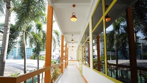 A corridor with a white floor, yellow and orange railings, and large windows with a view of palm trees and a road. - RoomQuest Hotel Pratunam (Bangkok)