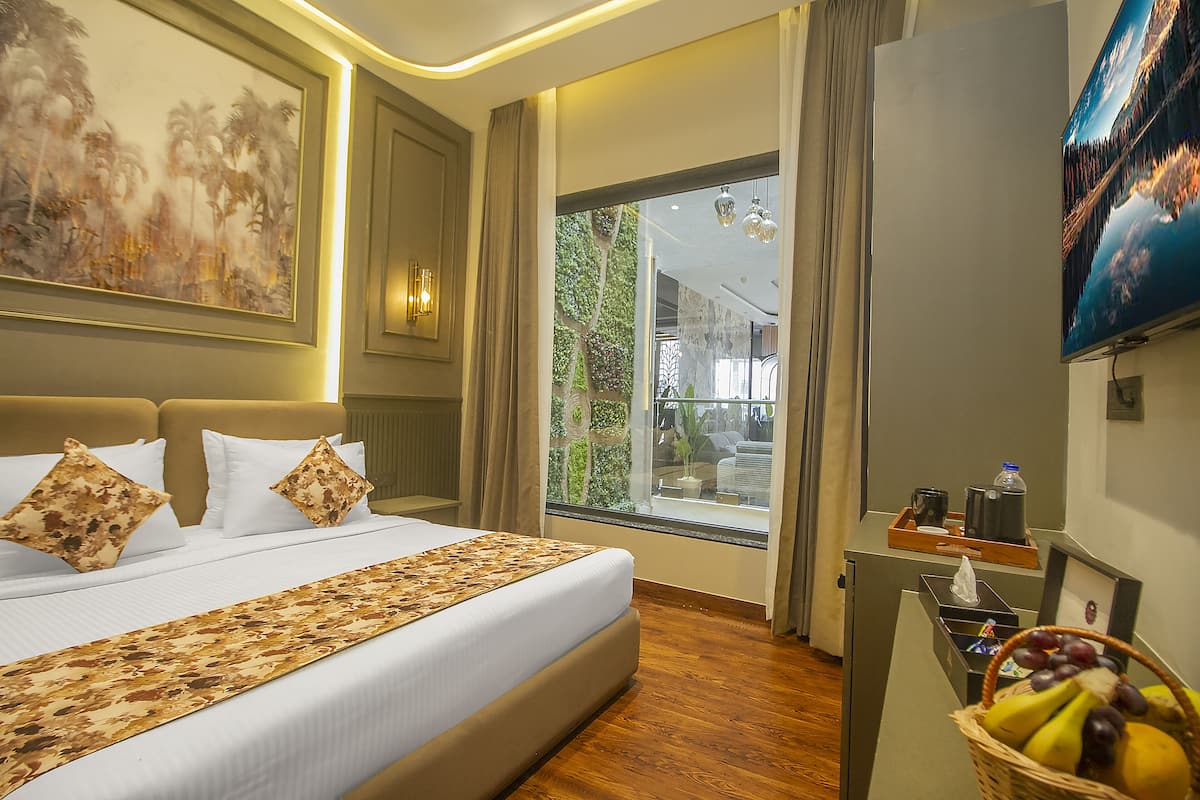 A hotel room with a large bed, a TV, a window with a view of greenery, and a small desk with a basket of fruit.