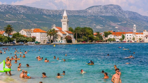 A crowded beach with people swimming and sunbathing, a prominent church with a bell tower, and mountains in the background. - Apartment Antonio with balcony, near the beach (Kaštel Novi)