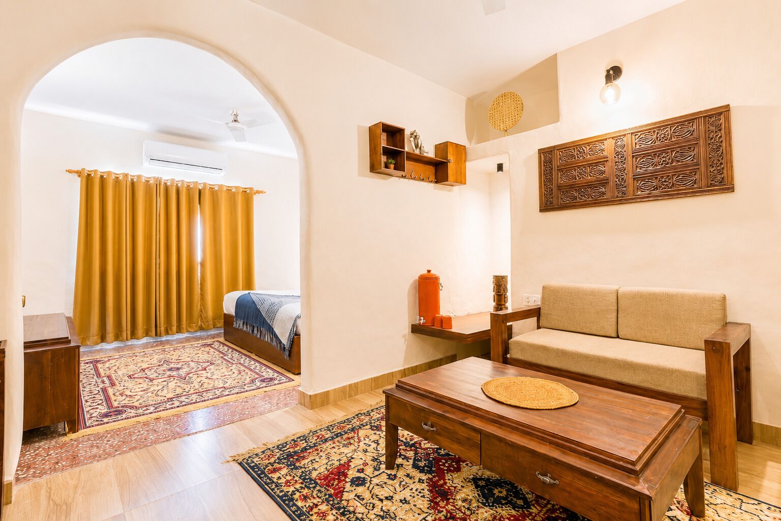 A hotel room with a bed, a sofa, a coffee table, a rug, a wall-mounted shelf, and an air conditioning unit.