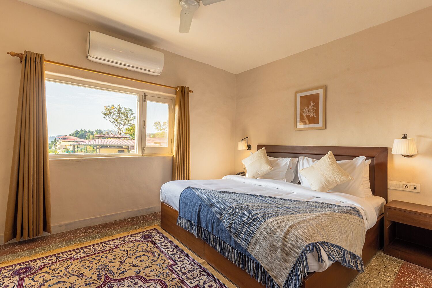 A hotel room with a bed, a window with curtains, an air conditioning unit, a nightstand with a lamp, and a patterned rug.