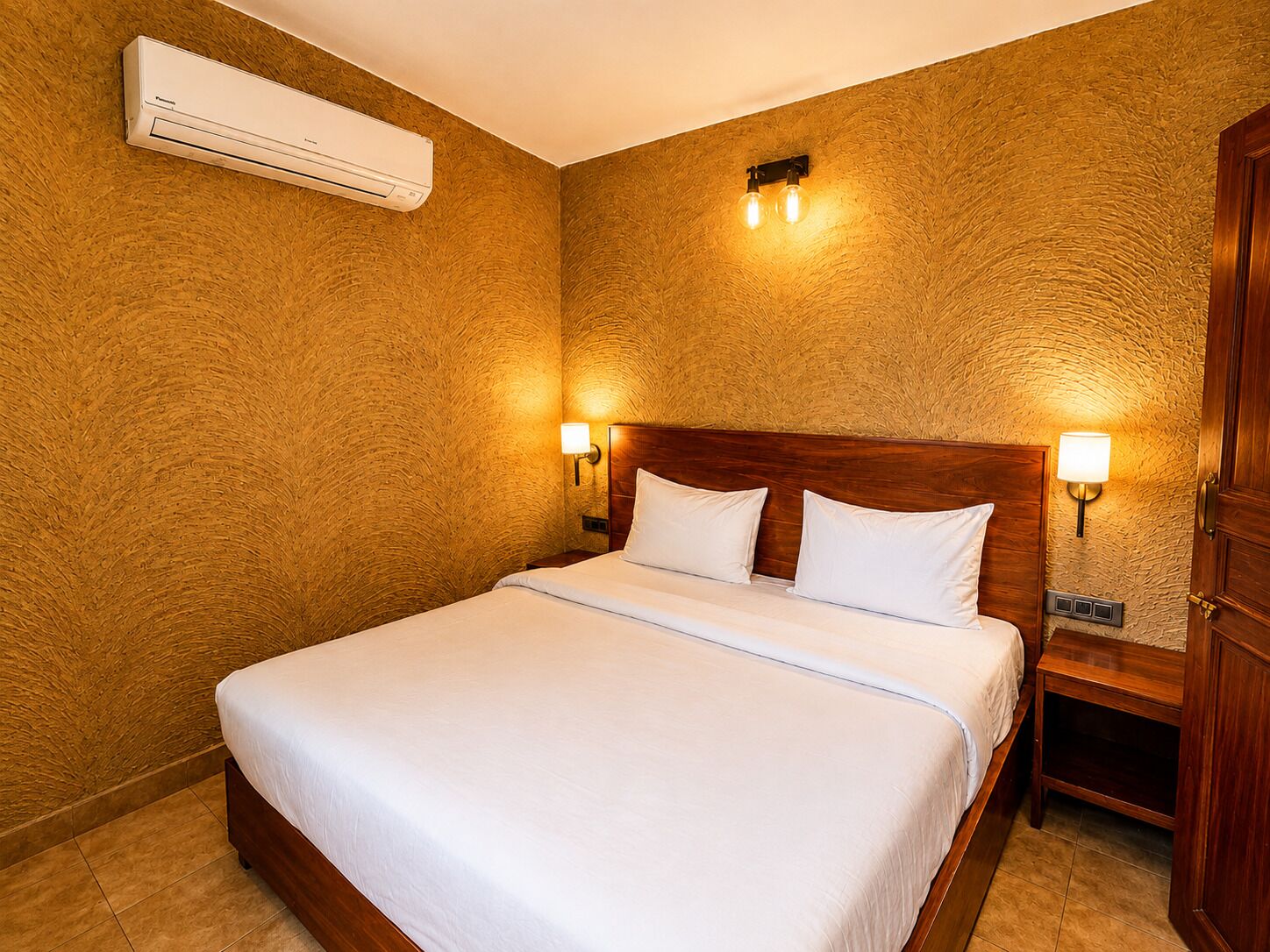 Habitación de hotel con cama de madera, ropa blanca de cama, aire acondicionado montado en la pared y dos lámparas de noche.