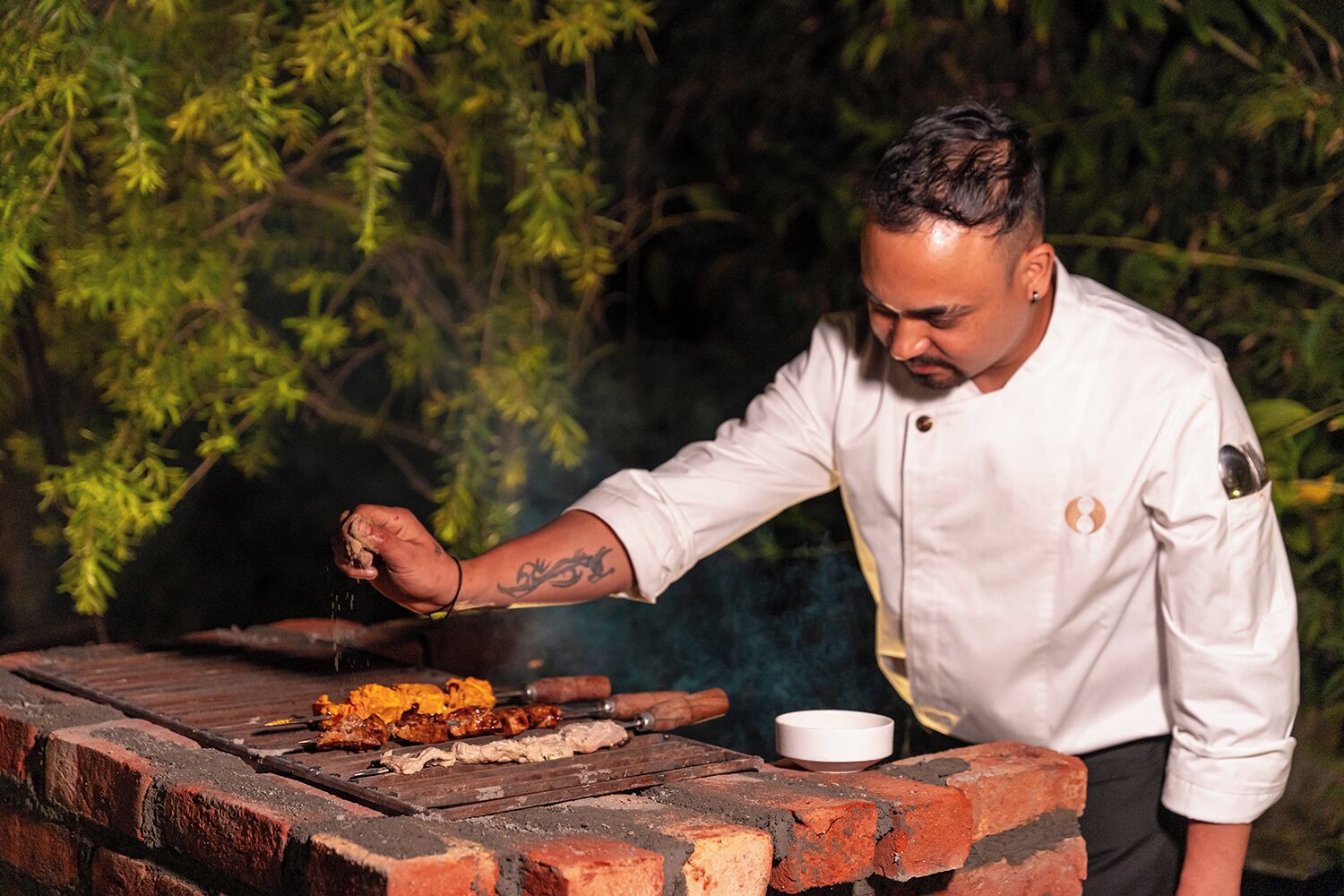 A chef grilling food outdoors with a brick grill and a pot.