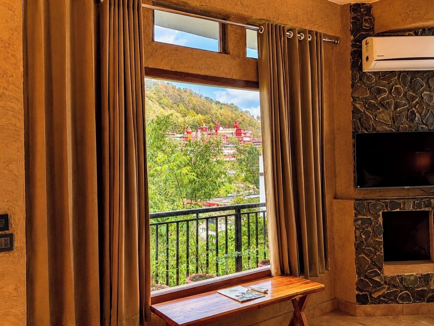 A room with a window overlooking a mountainous area, a stone wall, and a flat-screen TV mounted on the wall.