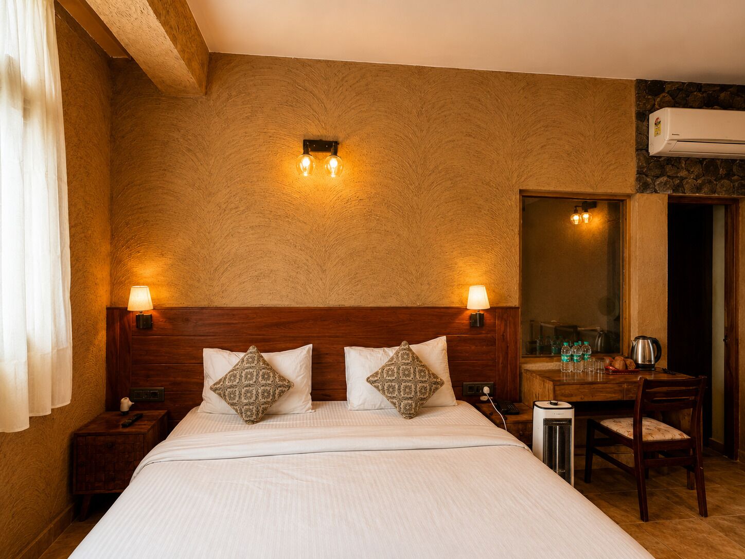 Ein Hotelzimmer mit einem großen Bett, zwei Nachttischlampen, einem Holzkopfschild, gemusterten Kissen, einem Nachttisch mit Fernbedienung, einem Schreibtisch mit Stuhl, einem Spiegel und einer Klimaanlage.