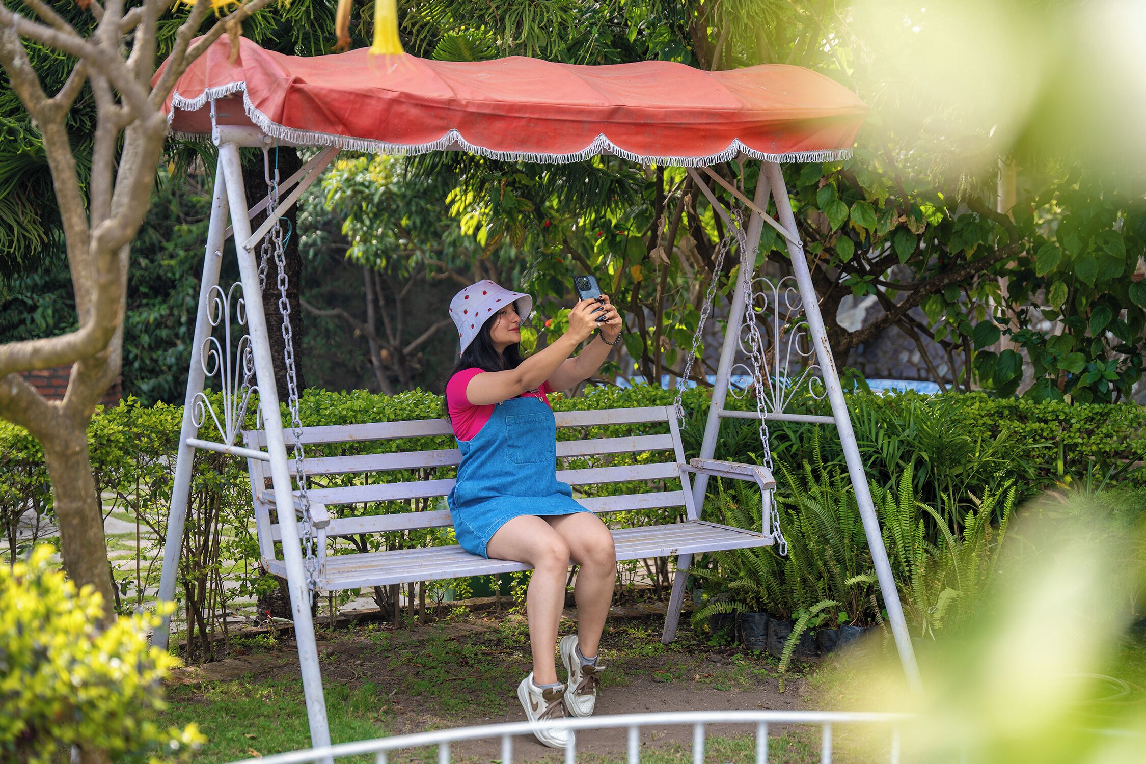 A person sitting on a white swing with a red canopy, surrounded by greenery.