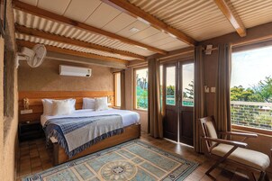 Un dormitorio con cama, mesita de noche y una silla. Hay una ventana con cortinas, un ventilador de techo y vistas al exterior con vegetación.