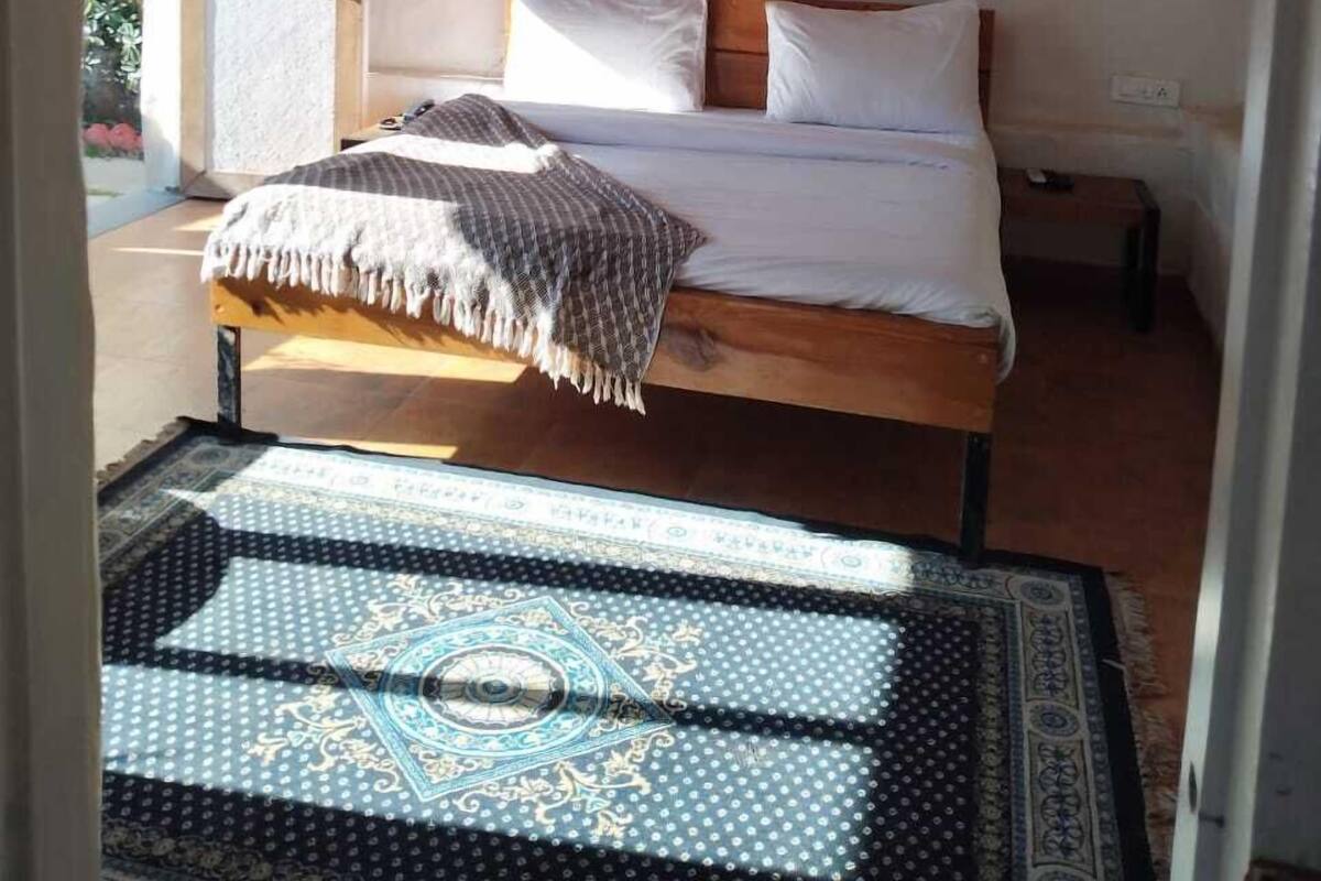 A single bed with a wooden frame, white bedding, and a patterned throw blanket. There is a ceiling fan, a framed artwork above the bed, and a small bedside table with a lamp.