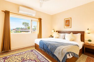 Quarto de hotel com cama, janela, ar condicionado, mesa de cabeceira, abajur e tapete.
