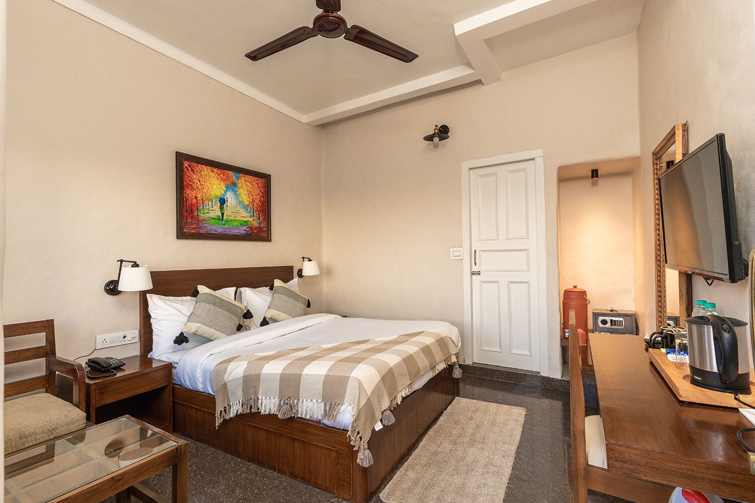 Habitación de hotel con cama, mesitas de noche, escritorio, silla, televisor, un cuadro, ventilador de techo y una puerta.