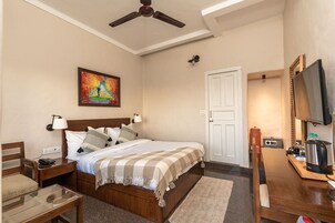Quarto de hotel com cama, mesas de cabeceira, escrivaninha, cadeira, televisão, pintura, ventoinha de teto e porta.
