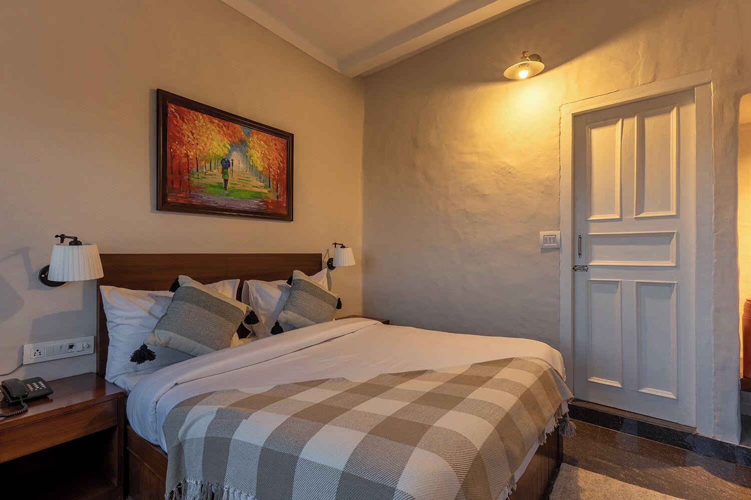 A neatly made bed with a checkered blanket, a painting on the wall, and a door to the right.