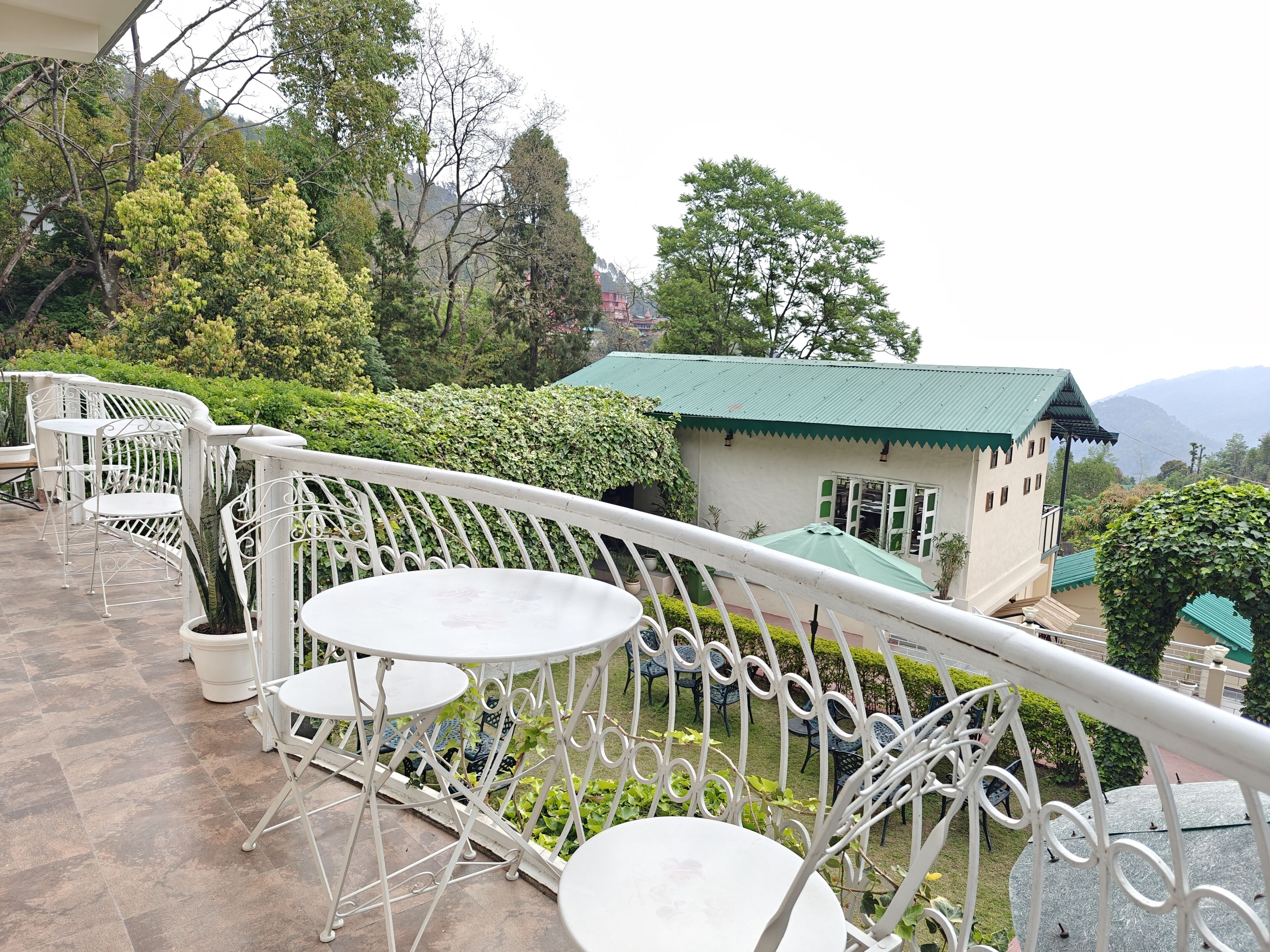 A terrace with white metal chairs and tables, a small building with a green roof, and surrounding greenery.