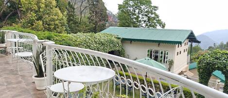 A terrace with white metal chairs and tables, a small building with a green roof, and surrounding greenery.