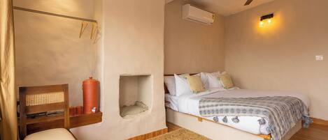 A bedroom with a bed, a chair, a small table, an air conditioning unit, and a ceiling fan.