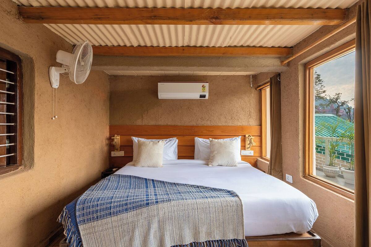 A single-bed room with a wooden headboard, a ceiling fan, an air conditioning unit, a window with a view of greenery, and a bedside table with a lamp.