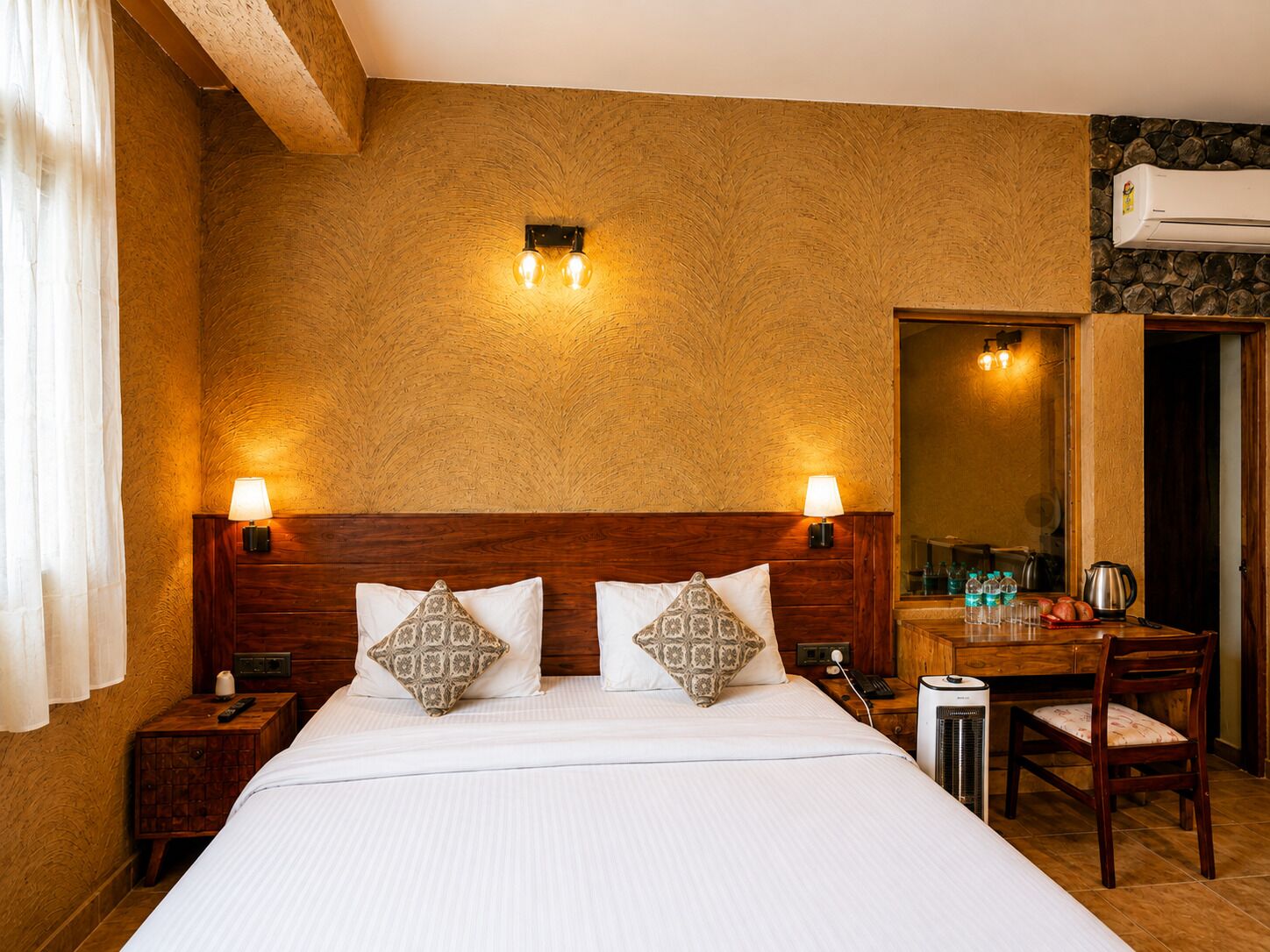 Hotelkamer met een groot bed, twee bedlampen, een houten hoofdbord, gestreepte kussens, een nachtkastje met een afstandsbediening, een klein kastje, een spiegel, een bureau met een stoel, een koffiezetapparaat en een airconditioning.