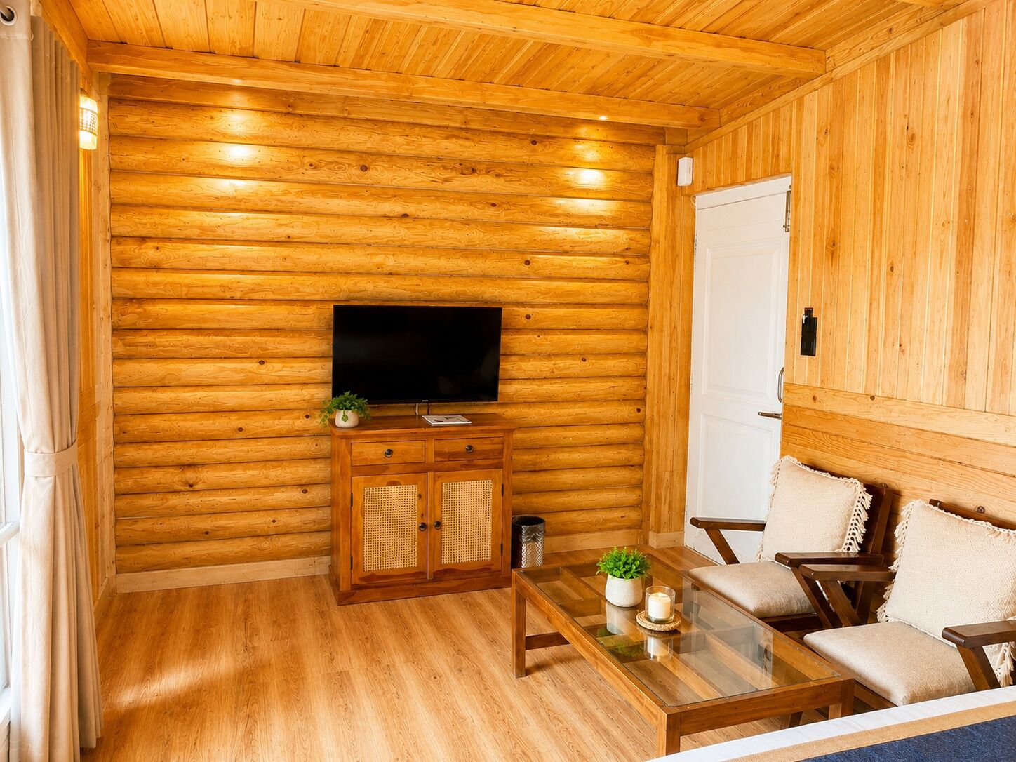 A wooden interior with a television on a wooden cabinet, a glass coffee table, and a sofa with cushions.