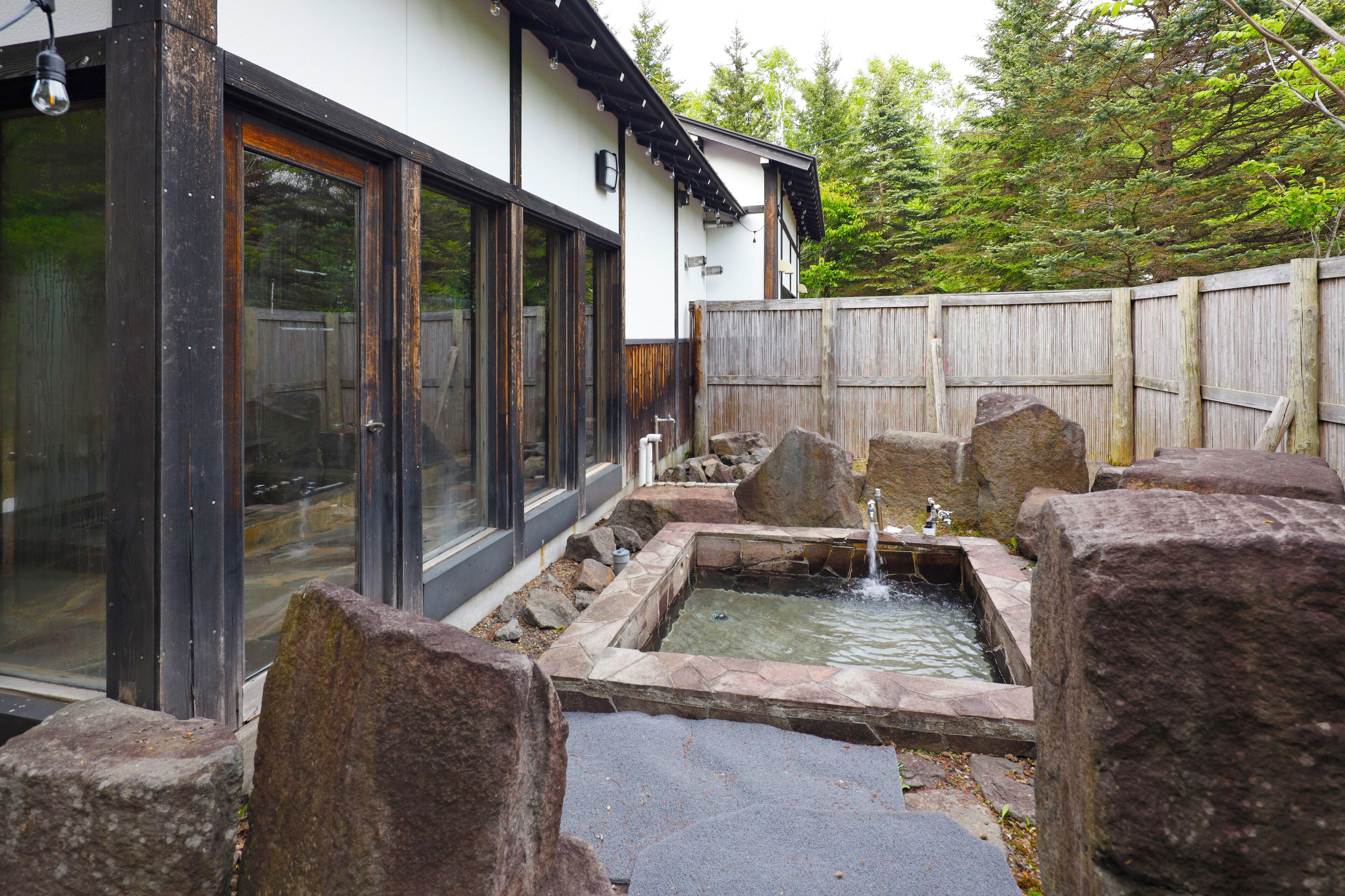 A traditional Japanese-style house with a wooden fence, a stone water feature, and a large window.