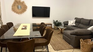 A modern living room with a glass dining table, brown cushioned chairs, a grey sofa, and a flat-screen TV mounted on the wall. - Casa Rural con Terraza y Barbacoa en Pleno Parque Natural (El Ojuelo)