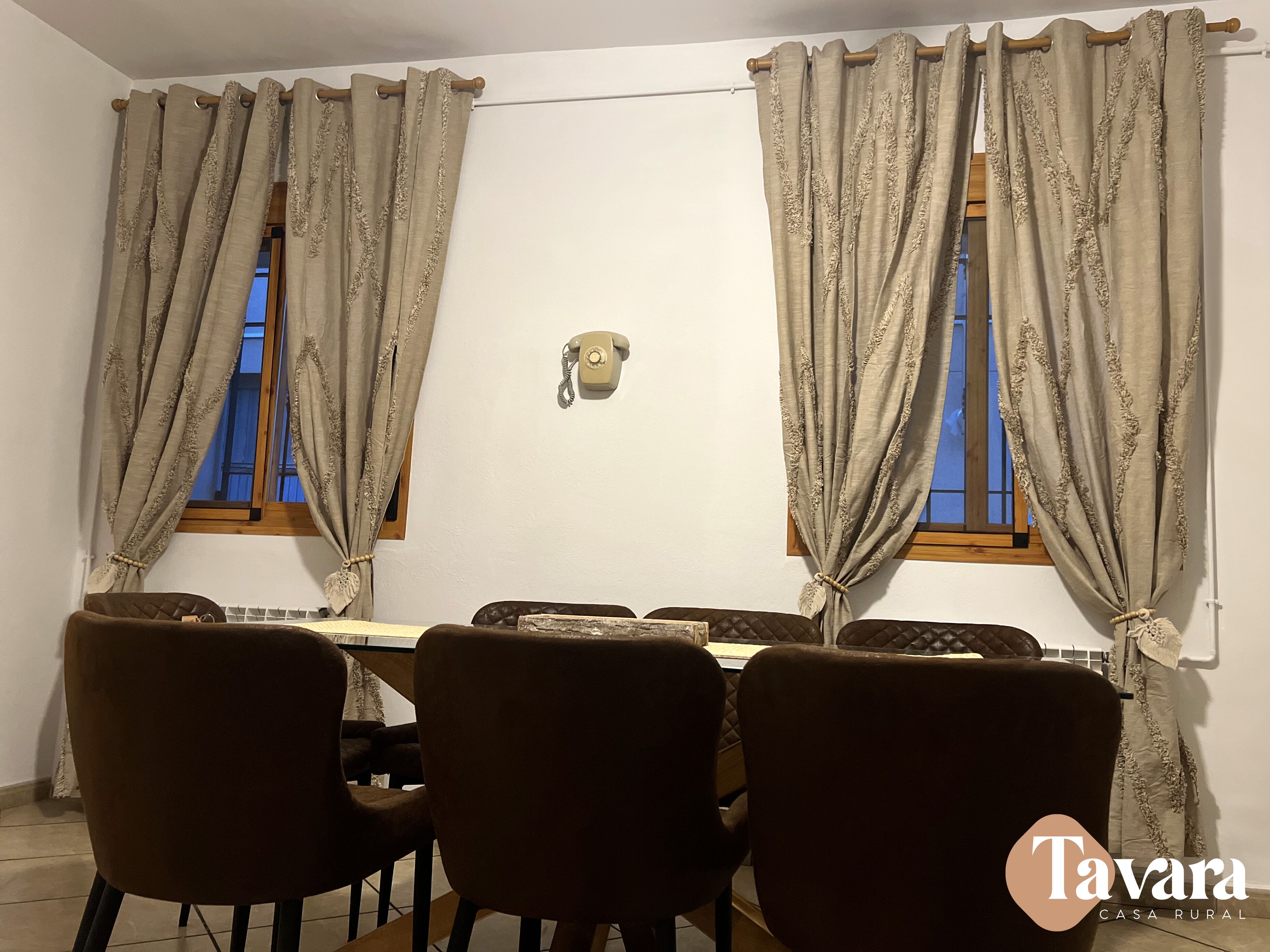 A dining area with a table set for four, two windows with curtains, a wall-mounted telephone, and a tiled floor.