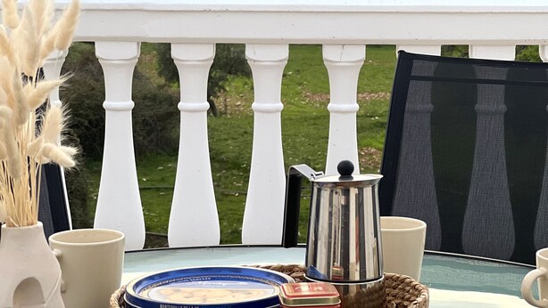 A balcony with a view of rolling hills, a white balustrade, and a table with a coffee pot, mugs, and a wicker basket containing chocolate and a tin. - Casa Rural con Terraza y Barbacoa en Pleno Parque Natural (El Ojuelo)