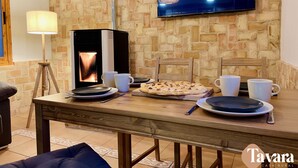 A cozy dining area with a brick wall, a flat-screen TV, a wood-burning stove, and a table set for two with plates, cups, and a pizza. - Casa Rural con Terraza y Barbacoa en Pleno Parque Natural (El Ojuelo)