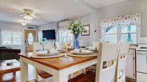 A dining area with a wooden table set for four, white chairs, a vase with flowers, and a ceiling fan. - Sand Life #1 by Bayley Vacation Rentals (Old Orchard Beach)