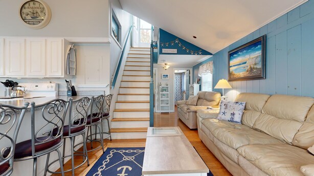 A living room with a beige sofa, a dining area with metal chairs, a staircase, and a kitchen with white cabinets. - Sand Life #2 by Bayley Vacation Rentals (Old Orchard Beach)